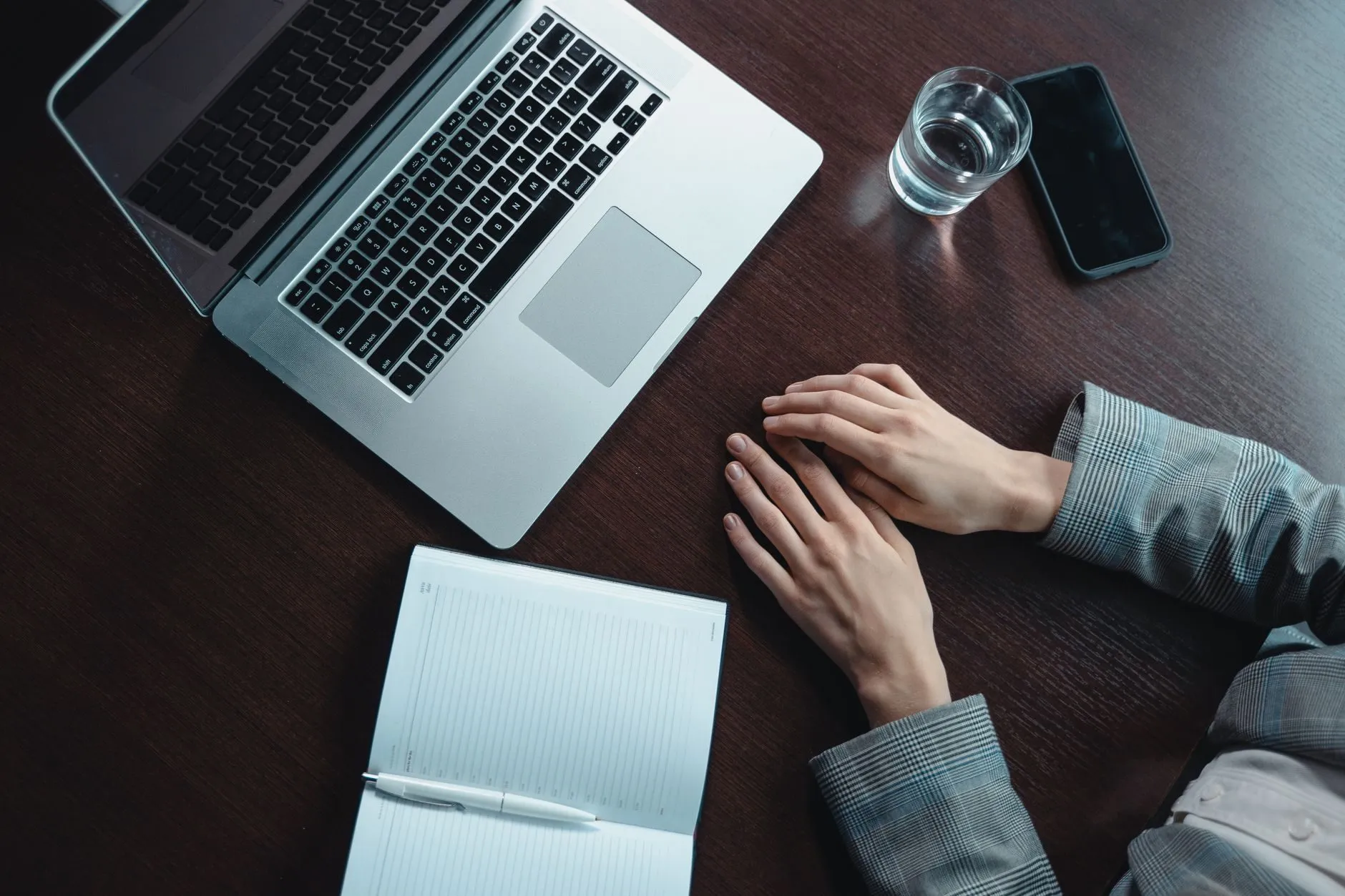 A laptop, an open notebook and a glass of water on a dark desk, hands resting beside them
