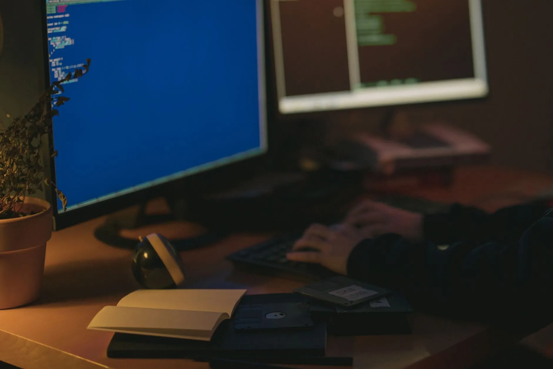 Two monitors with code on a desk at night, a hand on the keyboard in the background