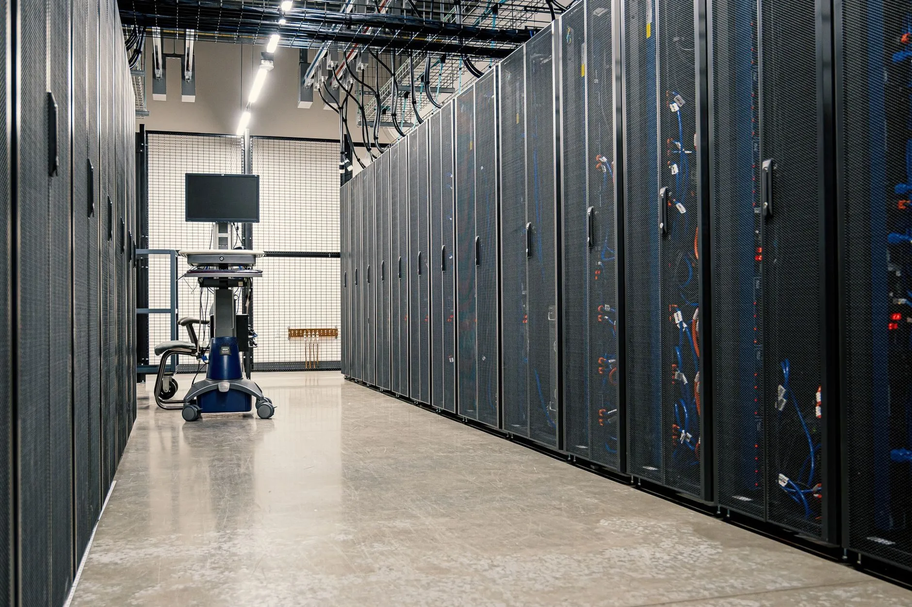 A server aisle with rows of black mesh-door cabinets and cables overhead