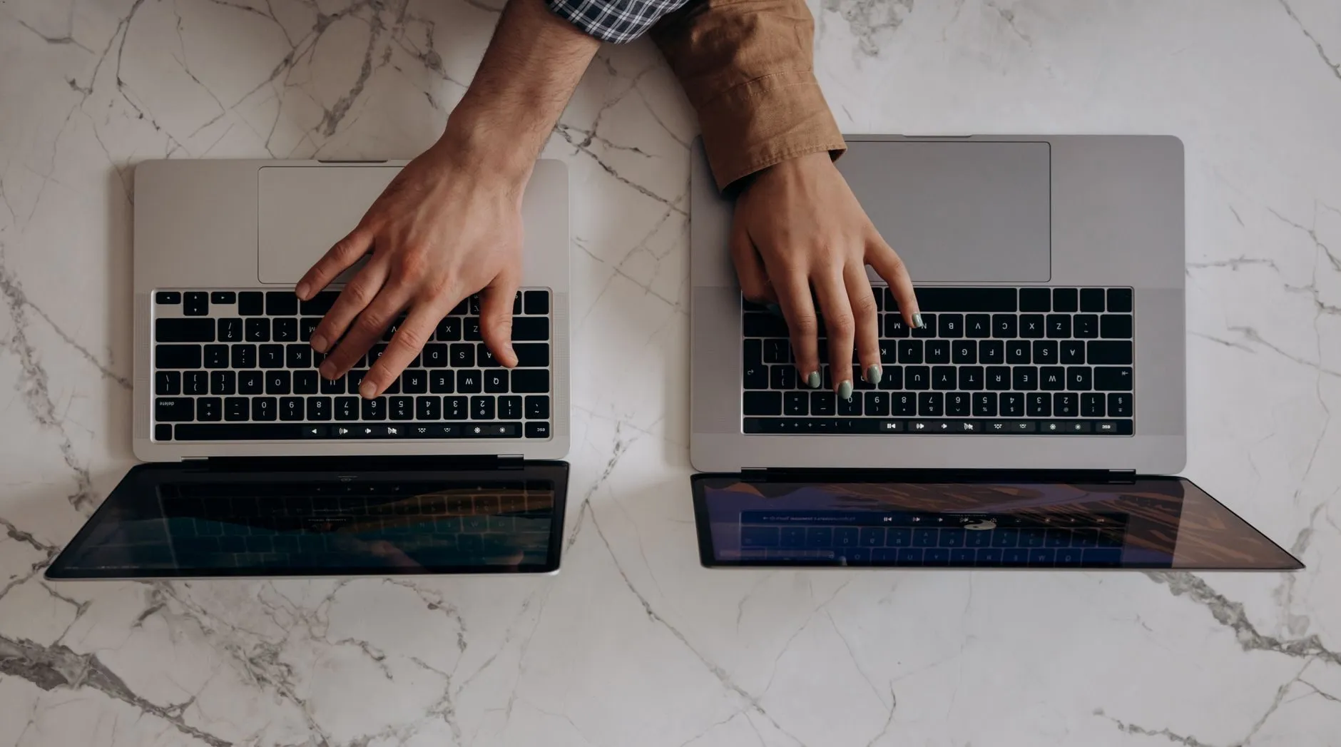 Two people’s hands typing on two laptops side by side on a marble table, seen from above